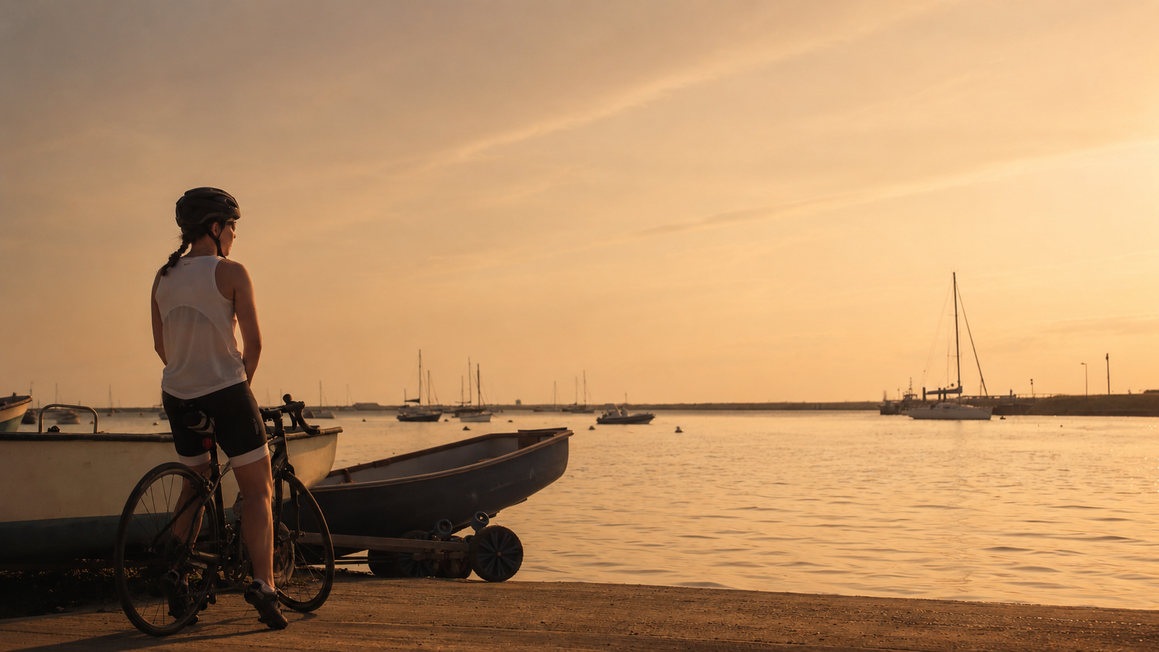 Cyclist pausing at the harbour's edge at golden hour, boats moored on the estuary